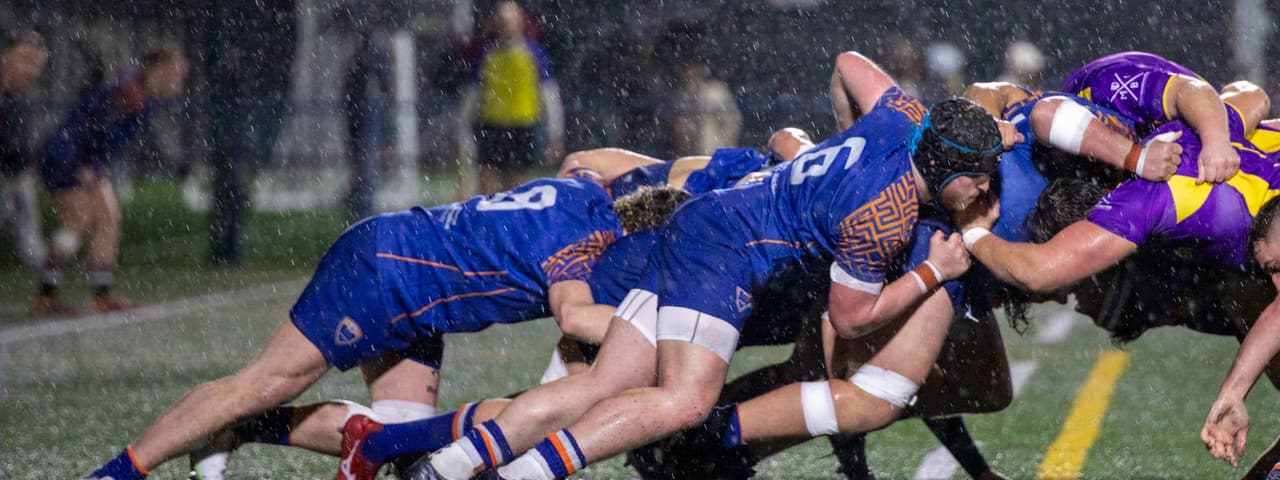 Two rugby packs locked in a scrum under floodlights during heavy rain